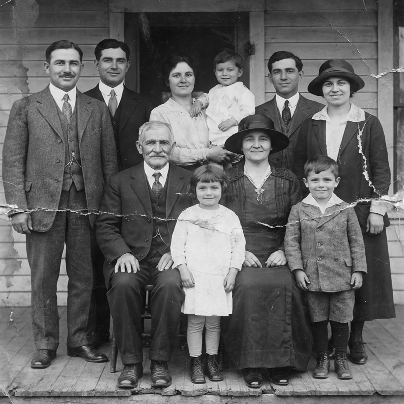 Black‑and‑white family portrait of eleven people posed in front of a wooden house, including adults and children dressed in early 20th‑century clothing.