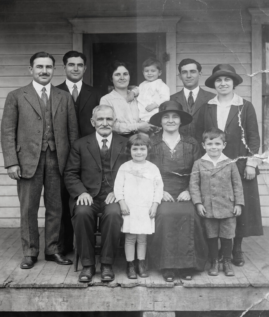 Black‑and‑white family portrait of eleven people posed on the porch of a wooden house, including adults in early 20th‑century attire and children in period clothing.