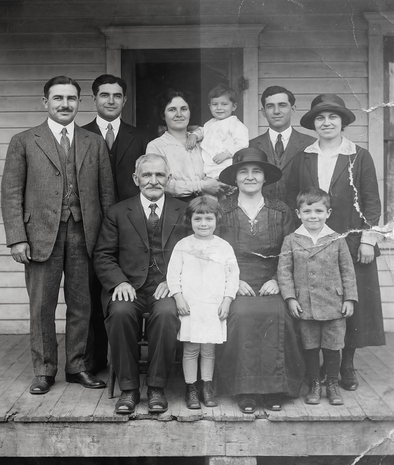 Black‑and‑white family portrait of eleven people posed on the porch of a wooden house, including adults in early 20th‑century attire and children in period clothing.