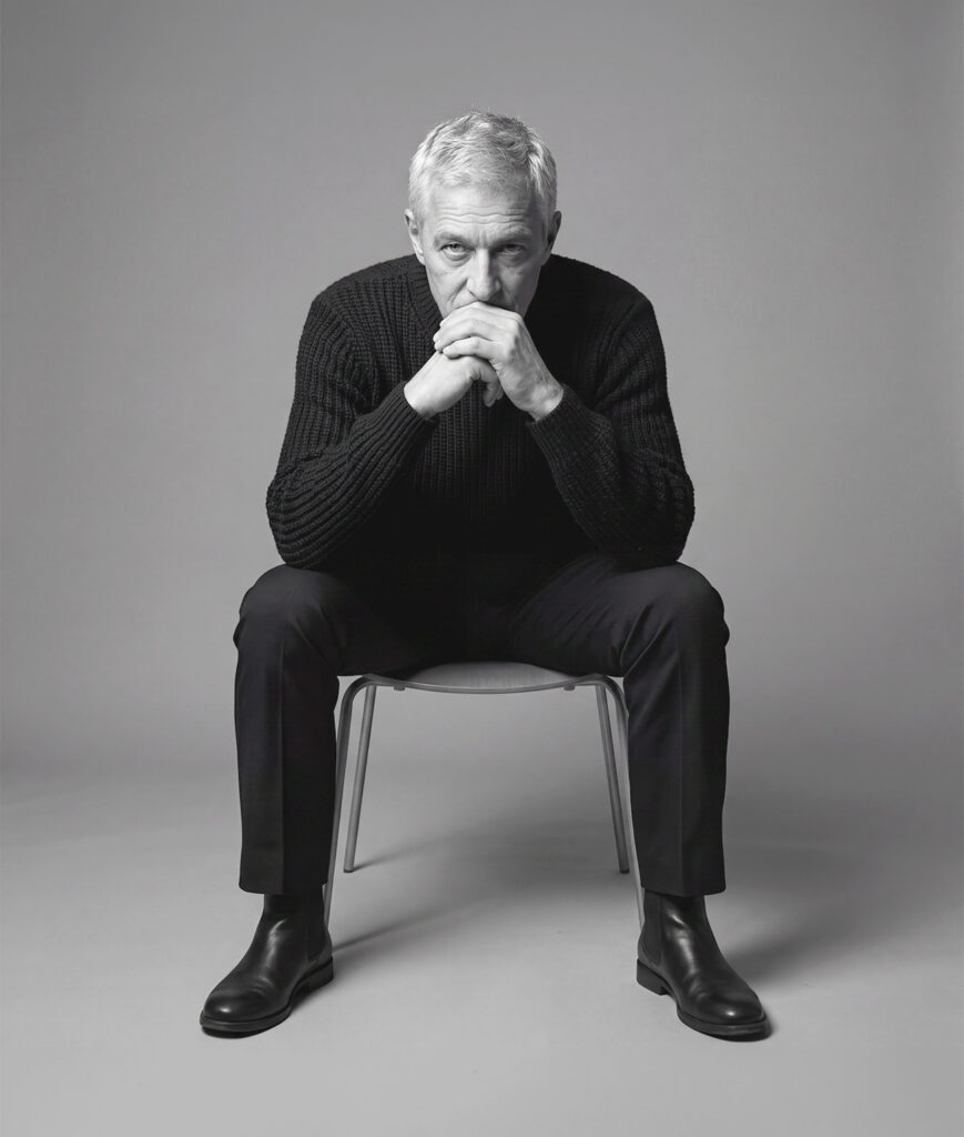 Black‑and‑white portrait of an older person sitting on a chair, leaning forward with hands clasped and looking directly at the camera against a plain background.